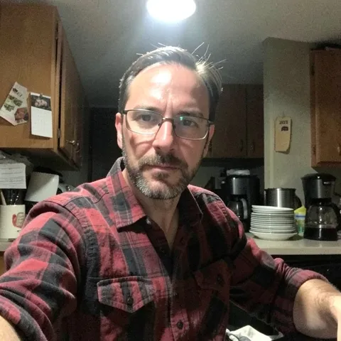 A bearded man in glasses and a flannel shirt in a dim kitchen at night.